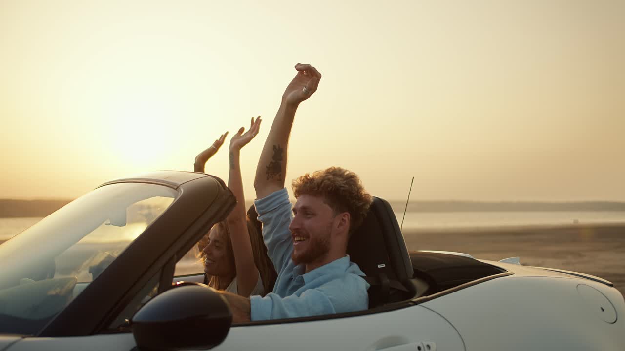 un tipo barbudo con cabello rizado en una camisa azul con su novia rubia están conduciendo en un descapotable blanco y agitando sus manos en un fondo de campo
