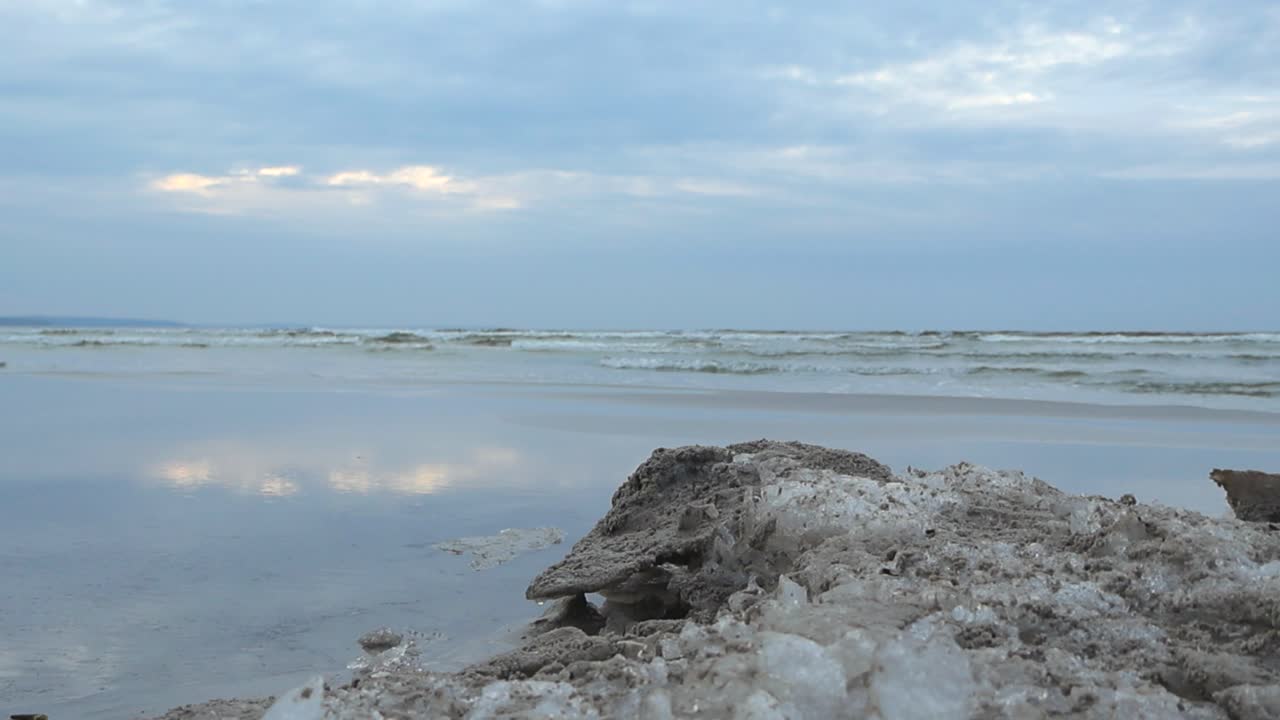 Low angle slow motion footage of a chunk of ice and snow on a reflective wet and damp sandy beach shoreline during a cloudy day while ocean sea water waves move with white water and foam in the back.