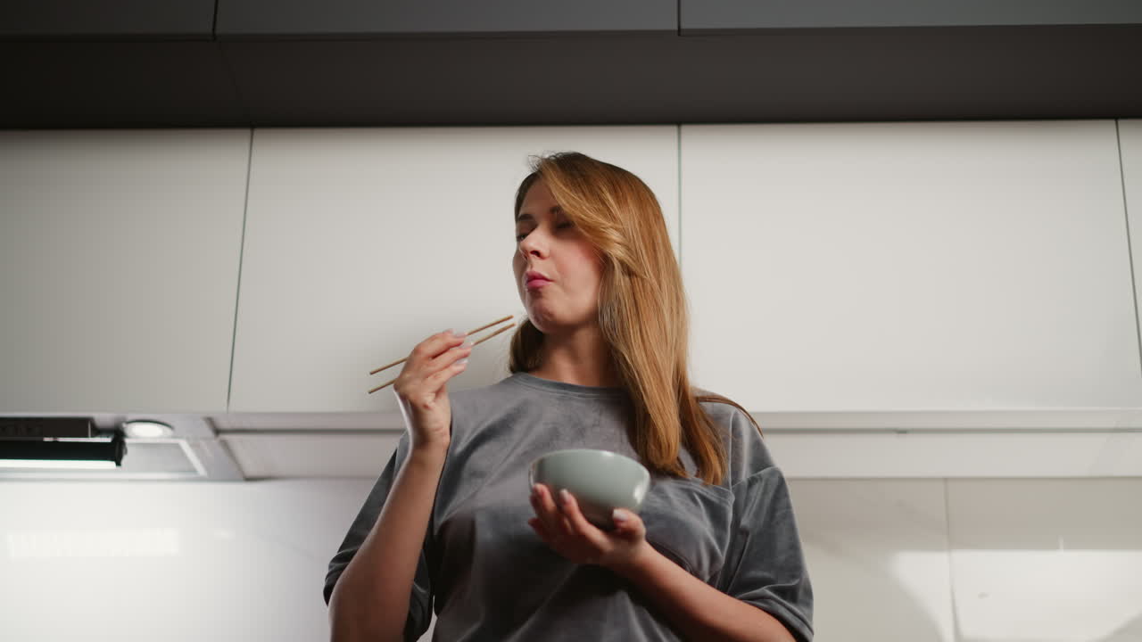 Young woman with long flowing hair stands in bright modern kitchen eating noodles with chopsticks while looking thoughtfully into background
