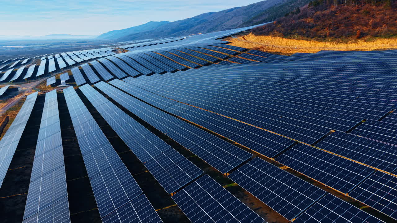 Luftfoto af rækker af solcellepaneler på en bjergskråning. Oversigtsbillede af en fotovoltaisk solcellepark installeret på bakket terræn under en blå himmel.