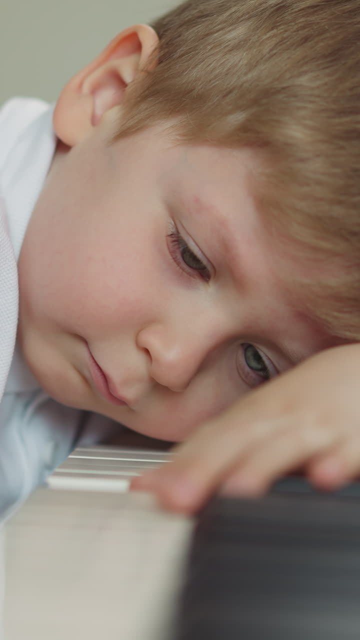 Child feels sleepy and sits with head resting on piano keyboard. Exhausted preschooler boy plays pressing key with finger. Learning music closeup