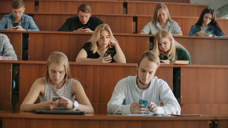 A group of men and women in a large university audience silently look at the screens of smartphones and write chat messages. Each person looks at the screens of his device without paying attention to the events of the real world.
