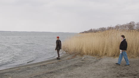 teenage boy dresses in an orange beanie and plaid coat standing near of seashore on a cloudy day, then a teenage girl in winter clothes approachs him