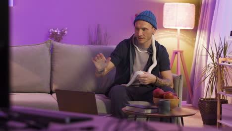 man sitting on sofa and holding a snake while watching something on the laptop