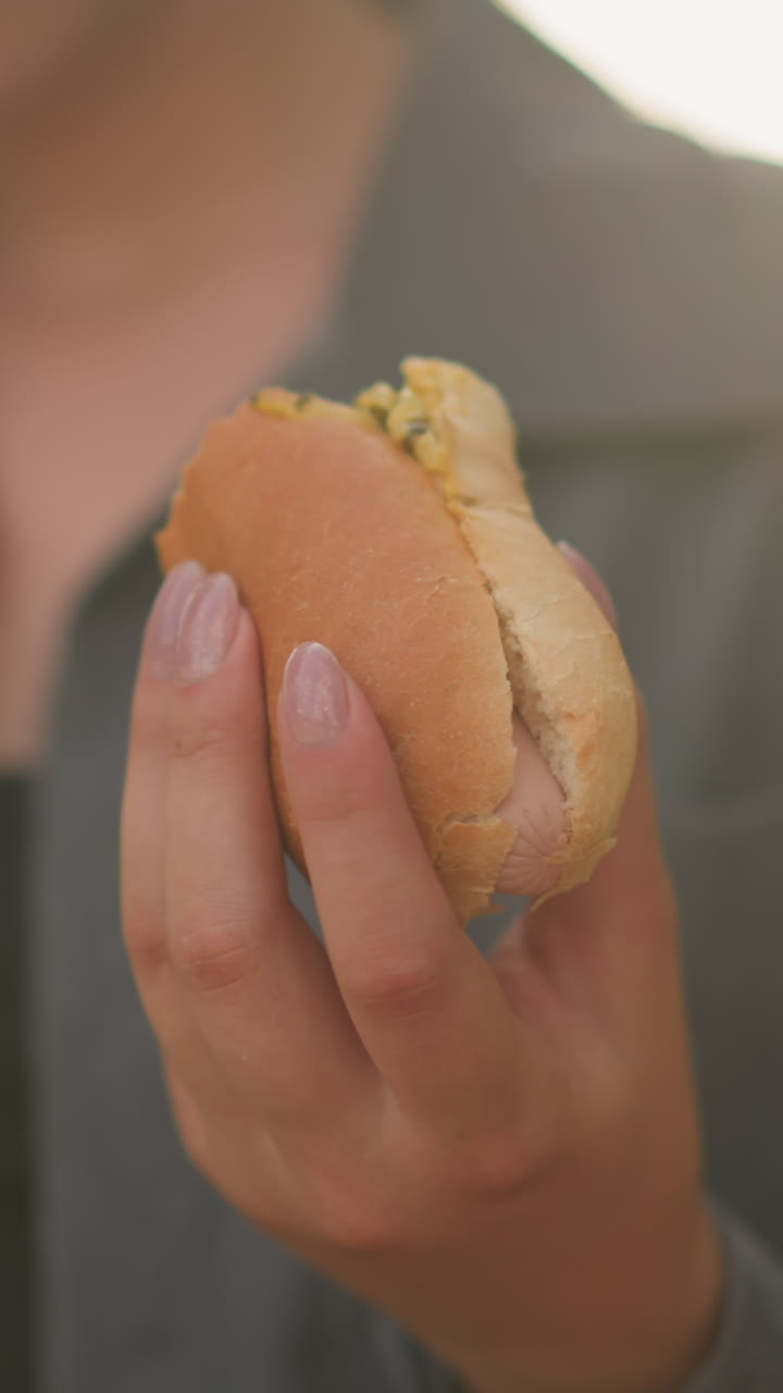 primer plano de una mujer con una camiseta gris sosteniendo shawarma mientras lee un libro en un entorno al aire libre iluminado por el sol, la luz del sol suave ilumina su mano y el bocadillo, con un fondo borroso de vegetación y cálidos rayos de sol