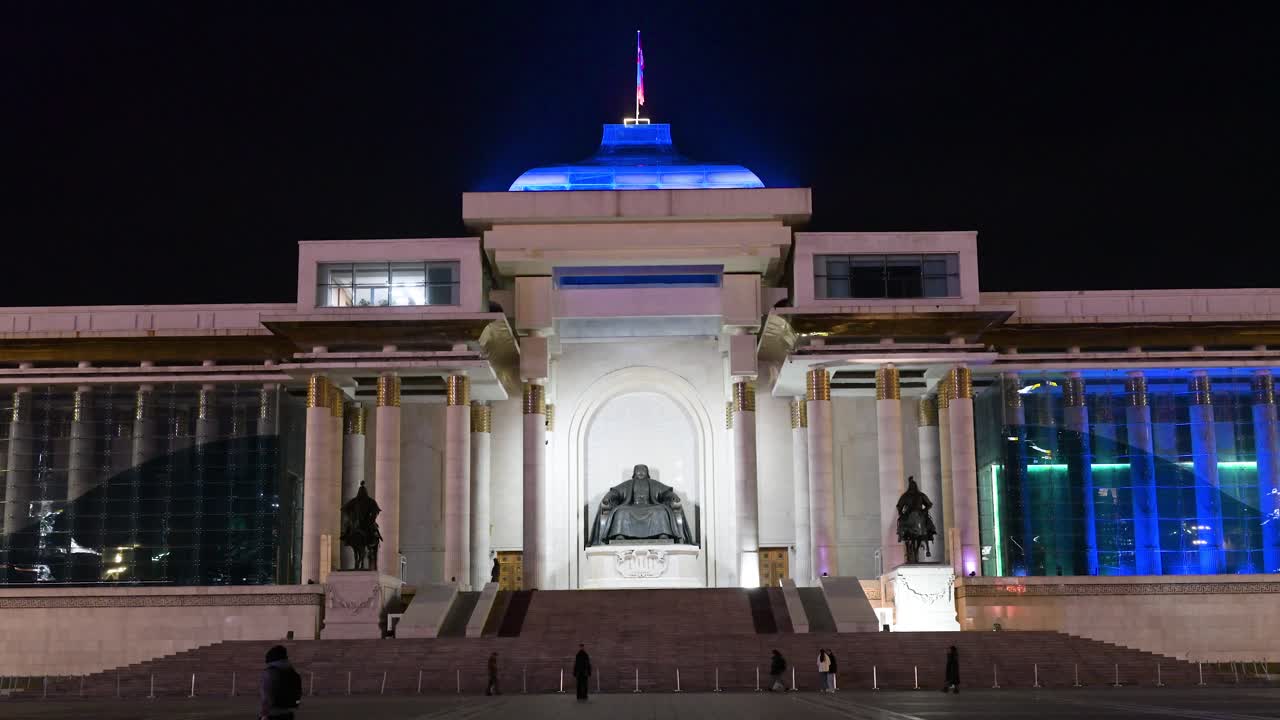The iconic bronze statue of Genghis Khan stands tall at Sukhbaatar Square, Ulaanbaatar, as people explore the illuminated space at night under the Mongolian flag.