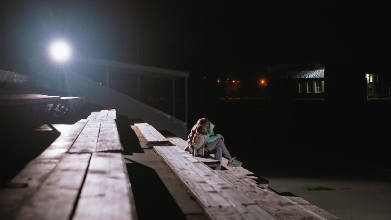 Wide view of dog owner sitting peacefully in park with puppy at night under bright spotlight, surrounded by empty benches and distant illuminated houses, capturing bond, and quiet companionship