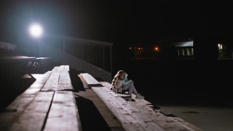 wide view of dog owner sitting peacefully in park with puppy at night under bright spotlight, surrounded by empty benches and distant illuminated houses, capturing bond, and quiet companionship