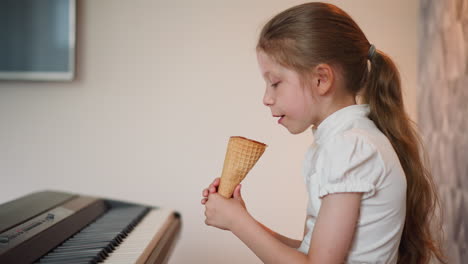 side view of little girl with long brown ponytail taking ice cream facing keyboard, wearing short-sleeve white shirt indoors, with calm focused expression against soft patterned wall background