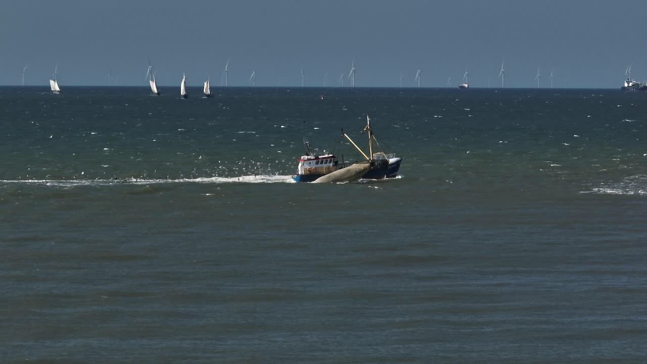 Side aerial view of fishing trawler boat followed by big flock of seagulls, boats sailing in background and wind turbines on horizon