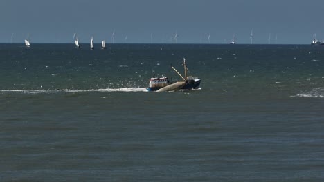 side aerial view of fishing trawler boat followed by big flock of seagulls, boats sailing in background and wind turbines on horizon