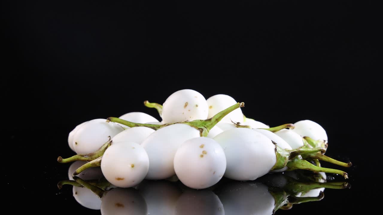 White Thai eggplants rotate smoothly on a reflective black surface, highlighting their glossy texture and vibrant green stems