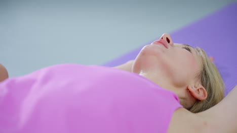 woman in pink lays back with arms raised, displaying a tired expression after exercise, slightly blurred background showcases fitness studio setting with equipment