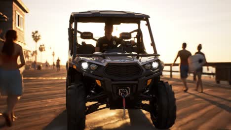 a police officer patrols on a utility vehicle along a beachside boardwalk during sunset. the warm glow highlights the vibrant atmosphere of the area with people enjoying the shoreline.