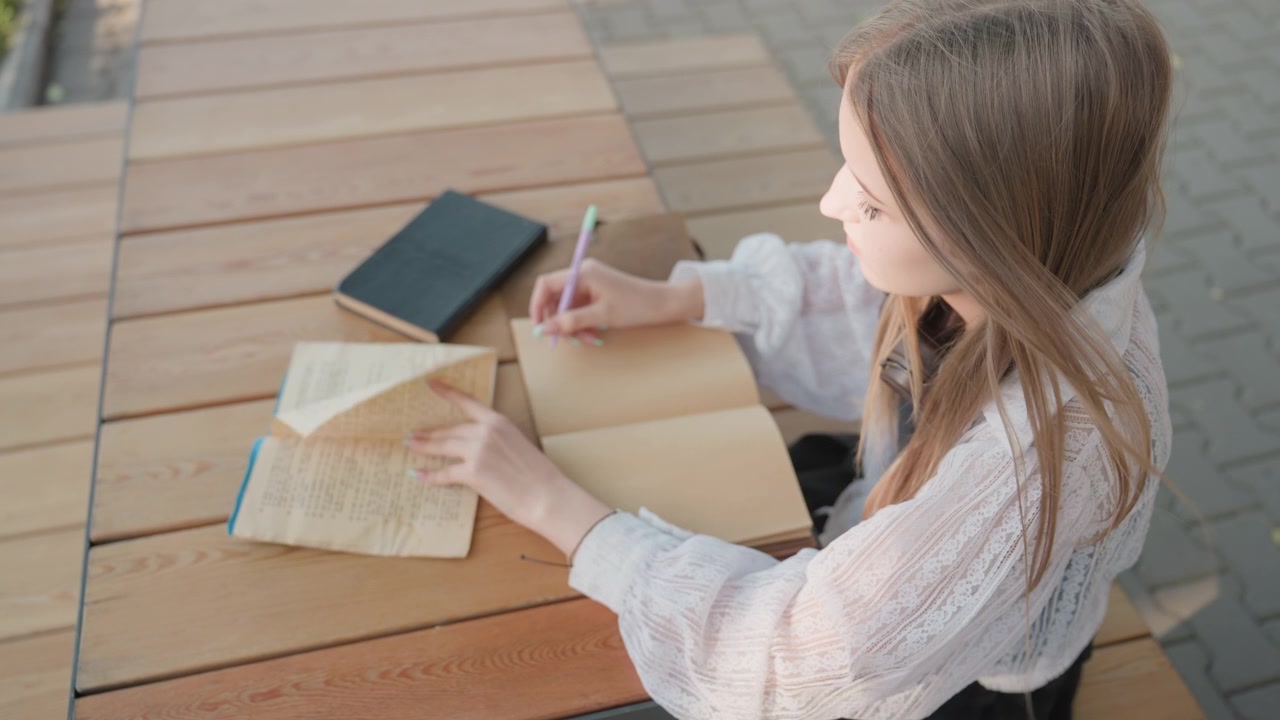 Close up of young woman reading old book and writing in notebook at wooden table outdoors. Woman wears white lace blouse interacts with open book pages using left hand while holding pen