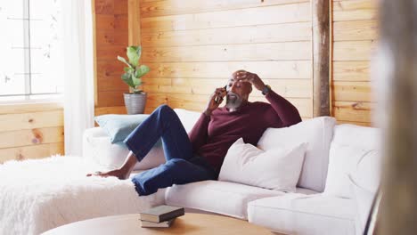hombre afroamericano mayor feliz pasando el tiempo en una cabaña de madera, sentado en el sofá y usando un teléfono inteligente