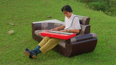 young asiatic artist playing a song on piano keyboard while sitting outdoor on a vintage couch recording a new album