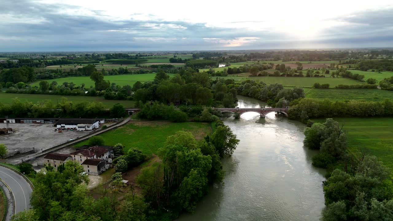 Un dron se desliza sobre el río Oglio en Pontevico, revelando un puente de arco de mampostería y la carretera que conecta Pontevico con Cremona, con el paisaje circundante de fondo.
