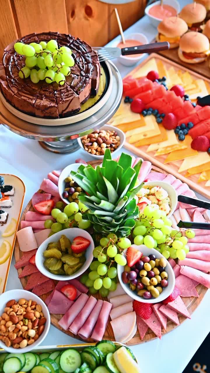 Вид сверху на разнообразные фуршетные блюда и мясное ассорти. Вид сверху на шоколадный торт, фруктовые тарелки, сыр, ветчину, орехи и овощи на белом столе.