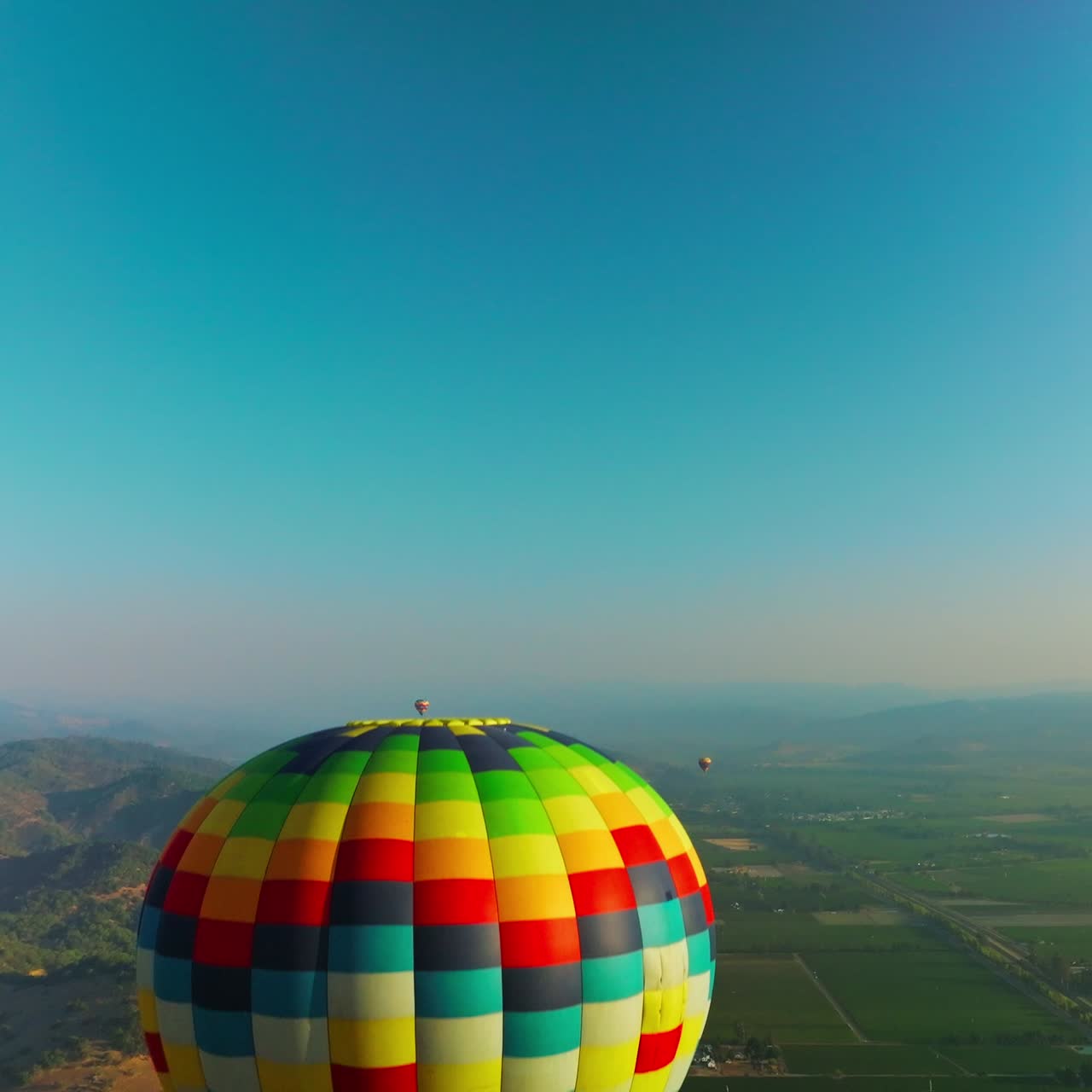Bright colorful hot air balloon descending in the blue sky. Aerostats soaring over the hazy mountains on sunny day. Top view