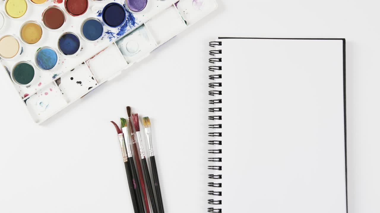 vista aérea del cuaderno, pinturas y pinceles sobre fondo blanco, en cámara lenta