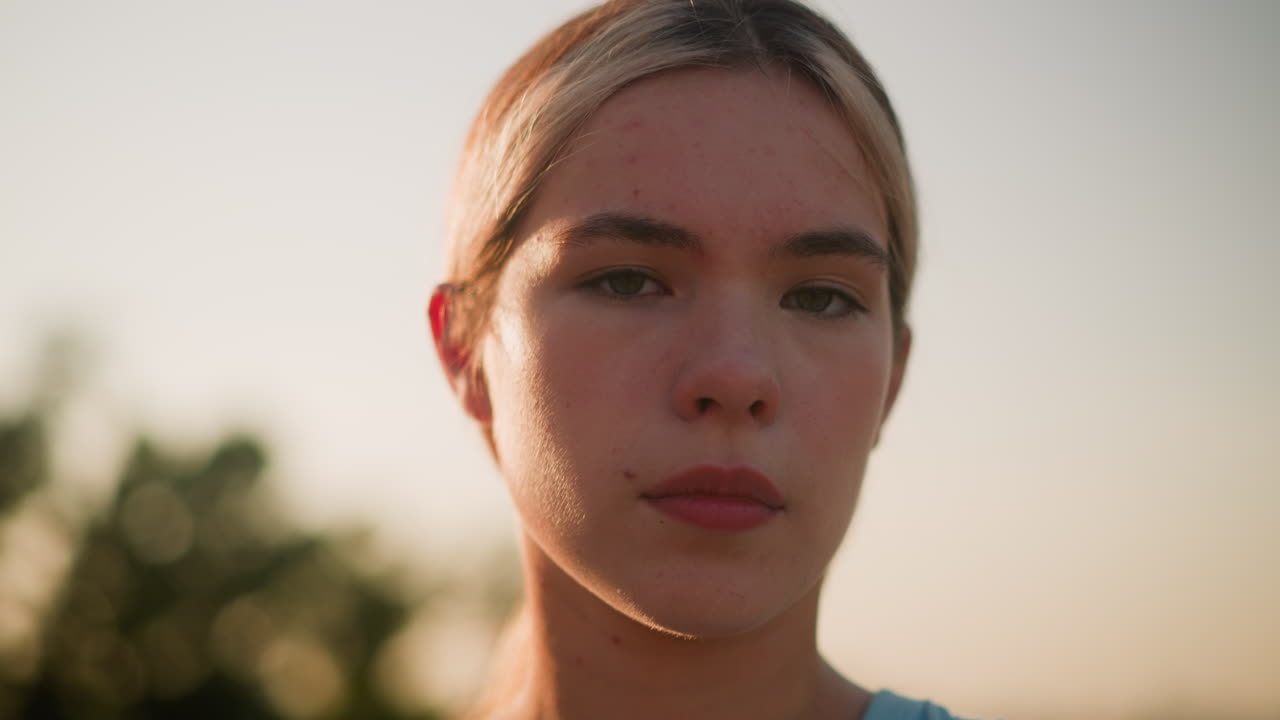 젊은 백인 여성의 클로즈업으로 기분이 좋지 않은 표정으로 카메라를 직접 바라보고, 따뜻한 빛 아래 눈을 이고, 배경에는 자연스러운 보케 효과가 있습니다.