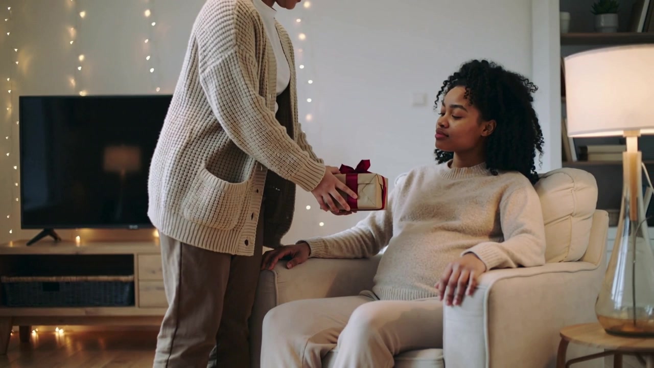 Partner che si avvicina tenendo un regalo incartato, posizionando il presente su una donna incinta in casa, per una festività