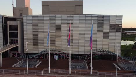 Drone-push-into-waving-flags-in-front-of-central-building-at-sunset-with-the-reflection-of-a-lake-in-the-background---Parque-del-Conocimiento,-Posadas,-Argentina