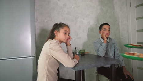 children sitting at kitchen table, one looking bored while other eagerly reaching out for sandwich, modern kitchen with sliced bread, condiments, and drinks on table for breakfast time