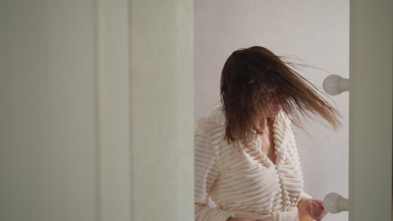 화장실에서 샤워를 한 후 머리를 흔들며 춤을 추는 여성. 로브를 입은 긍정적 인 여성은 램프와 함께 조명 된 화장실에 은 날아가는 머리카락을 회전시니다. 느린 동작