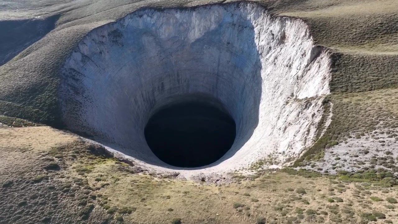 Luftfoto af massivt krater med stejl hvid kant og dybt mørkt centrum, solbeskinnede græsbevoksede skråninger med erosionsar.