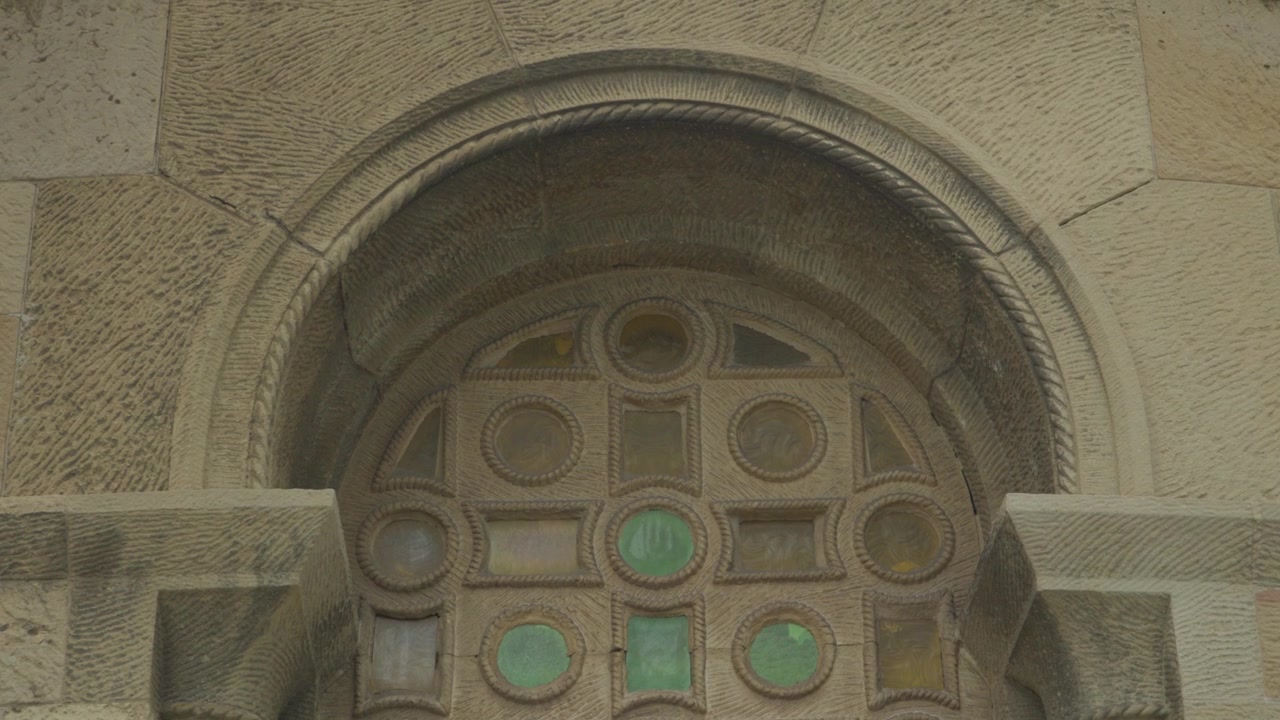 Close-up estático de um arco românico de pedra esculpida e janela geométrica decorativa com círculos de vidro verde na Igreja de San Pedro, Gijón.