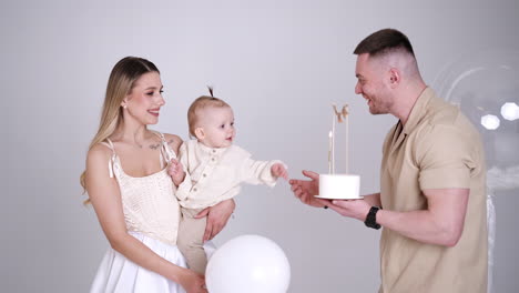 blonde woman holds a baby in hands. kid turns to a man approaching with a cake. celebrating first child's birthday.