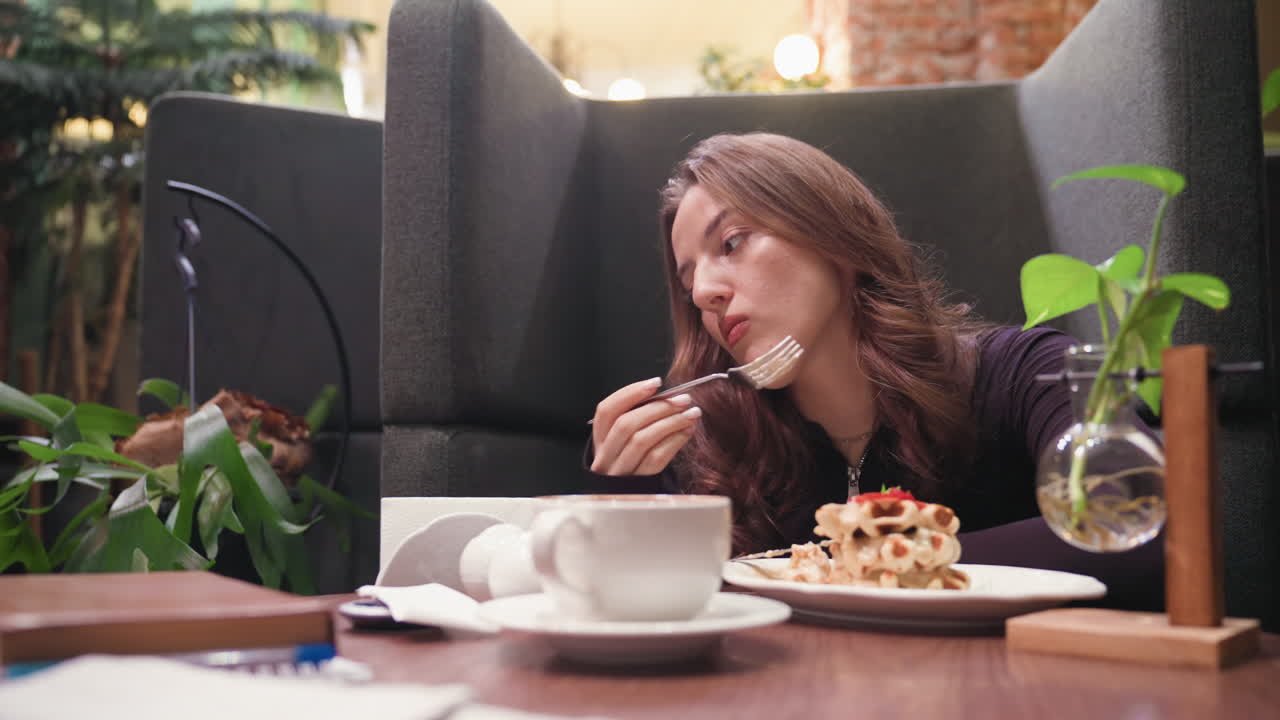 Lady sits in cozy booth holding waffle with contemplative expression, surrounded by warm and soft lighting, white cup and plant vase on wooden table creating calm reflective atmosphere