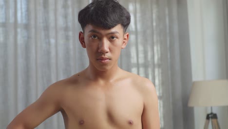 아시안 십대 소년이 집에서 셔츠를 벗고 운동한 후 카메라에 미소 짓고 있다.