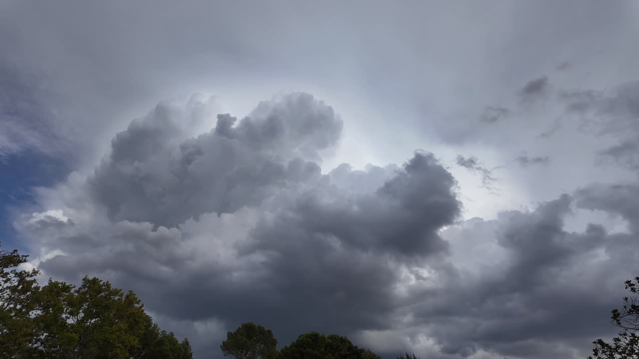 Timelapse of a threatening and massive storm cloud evolving in a turbulent blue sky over some trees. Daylight