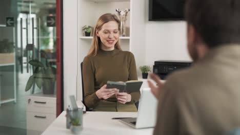 two colleagues are sitting next from each other and talking;they are engaged in conversation,discussing a new successful project.the colleagues are in a modern office,and the woman is holding a diary