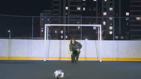 portero con capucha rodando una pelota de fútbol en el suelo durante la práctica nocturna en el campo al aire libre, área deportiva urbana cercada con luces de la ciudad en el fondo