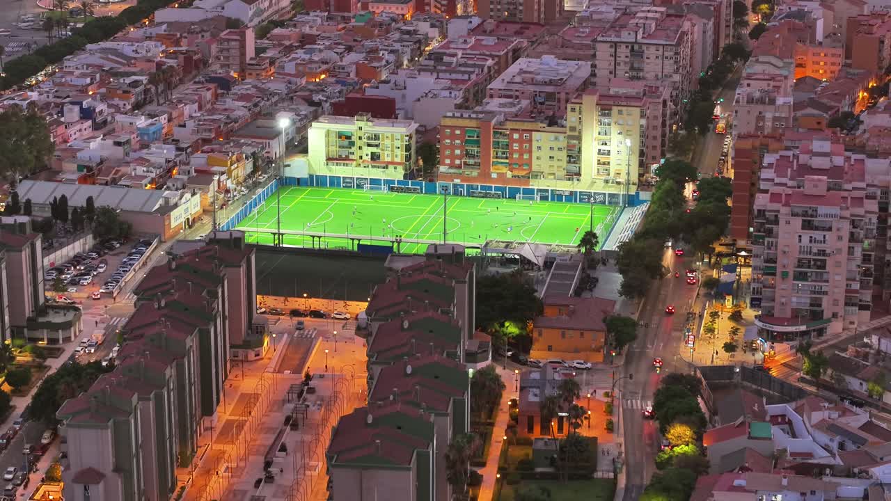 Aerial view over Málaga at dusk, revealing a brightly lit football field surrounded by dense urban housing and city streets glowing with evening lights