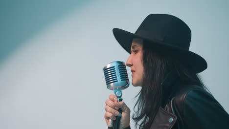 passionate female singer in a black hat and leather jacket, holding a vintage microphone with one hand and moving the other expressively, performing against a white background