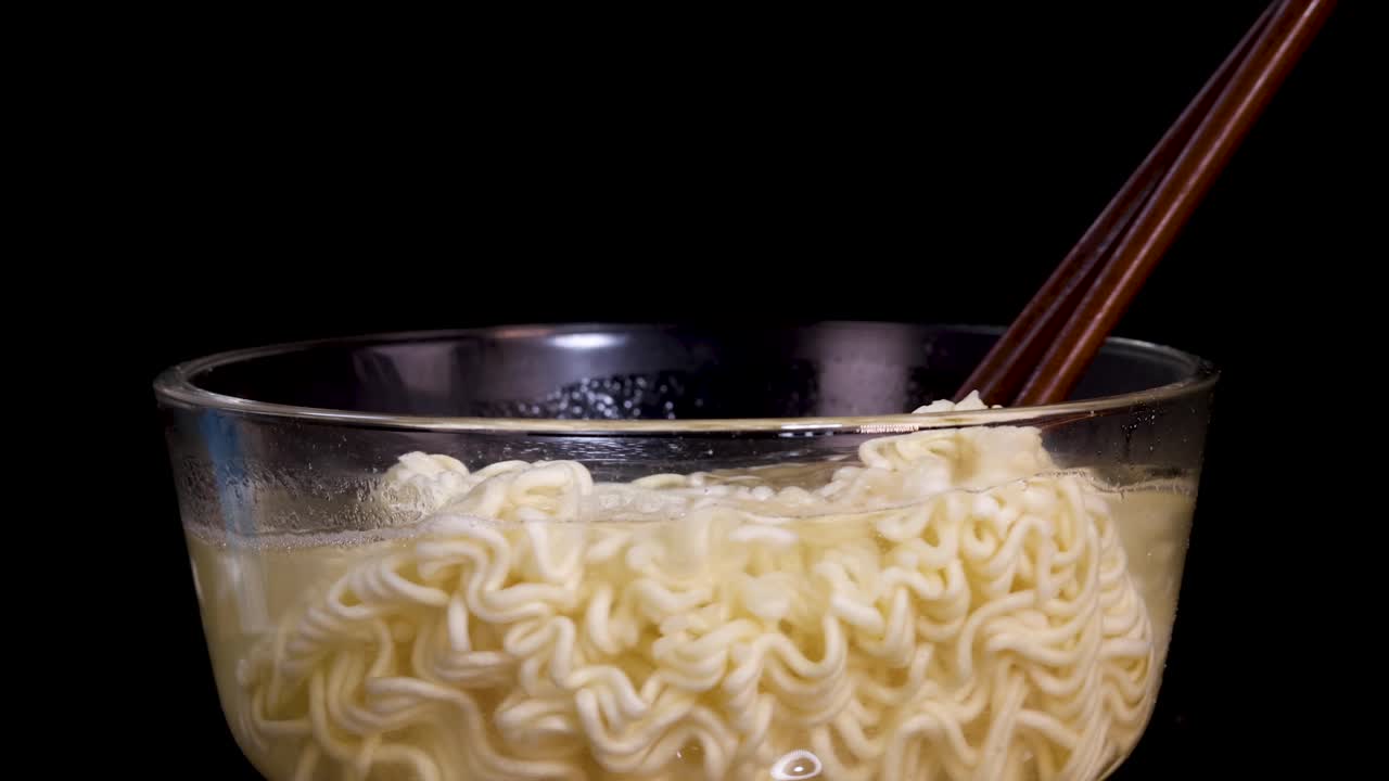 Chopsticks stir cooked instant noodles in a clear glass bowl against a black background, with steady camera and bright, even lighting highlighting the noodles’ texture