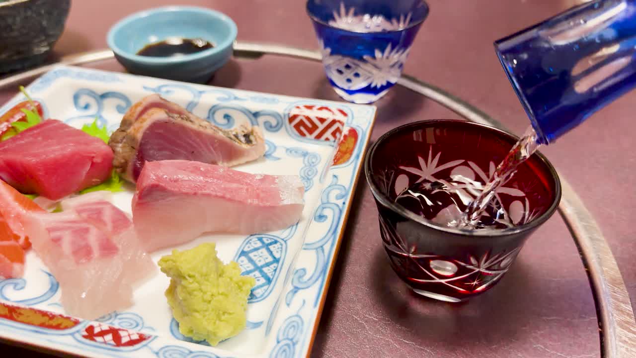 Klarer Sake wird in ein rotes Glas gegossen, neben einer Platte mit frischem rohem Fisch.