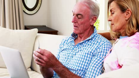 Smiling-senior-couple-doing-online-shopping-on-laptop-in-living-room