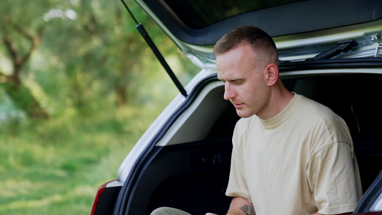 Focused male freelancer looks at the screen of his laptop. Man works remote sitting in the car trunk in forest.