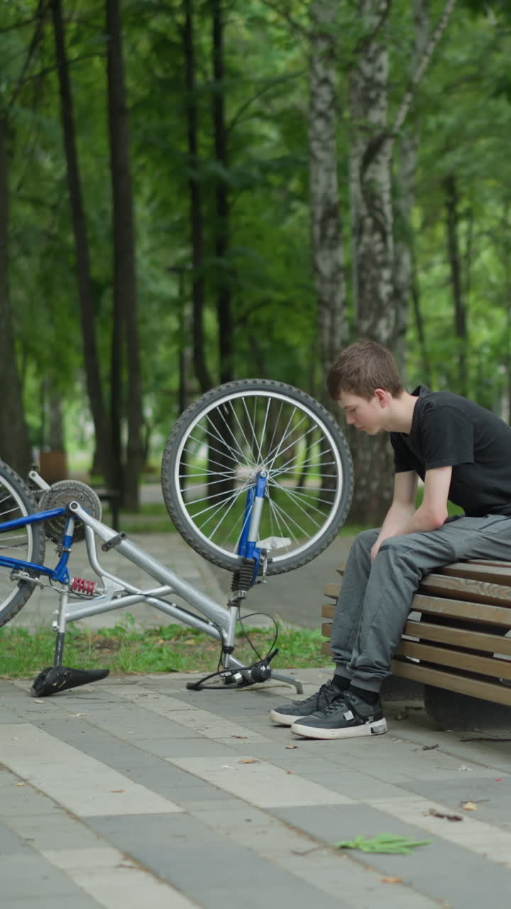 un niño se sienta en un banco del parque con la cabeza inclinada, su bicicleta colocada al revés a su lado, otro niño se acerca en bicicleta, y comienzan una conversación en un parque sereno