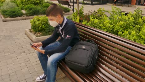 teenage boy wearing face mask approaches to the bench, sit down and play on the mobile phone