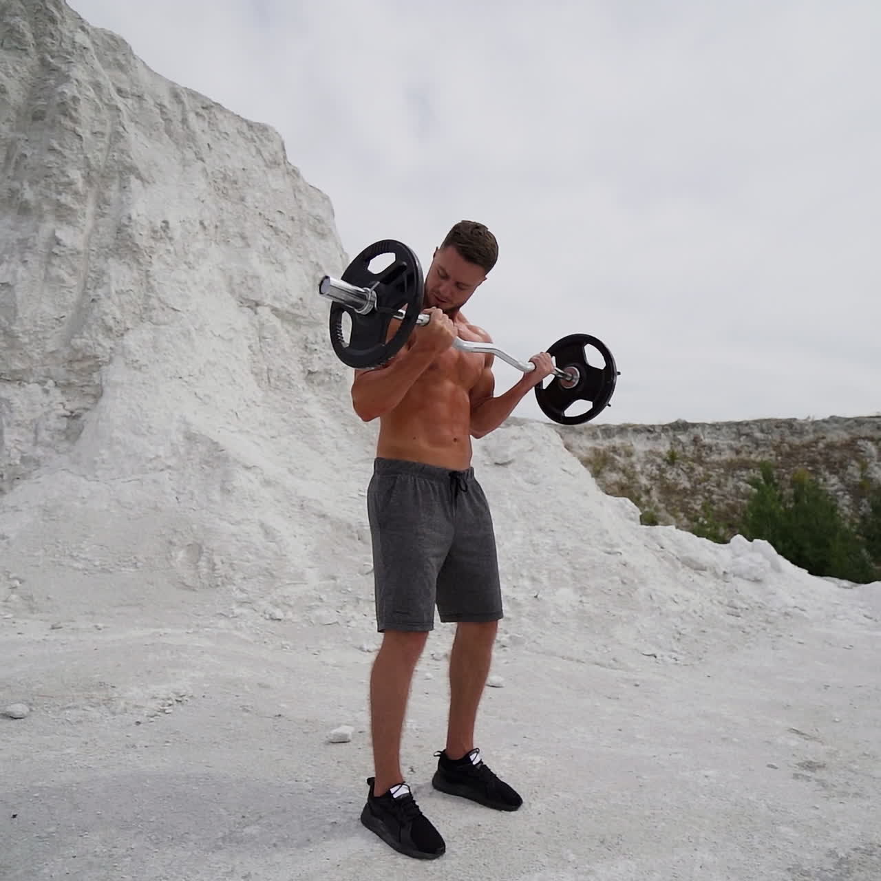 Handsome athlete training outdoors. Strong shirtless man with muscular body is lifting barbell on the white mountain background in summer. Slow motion.