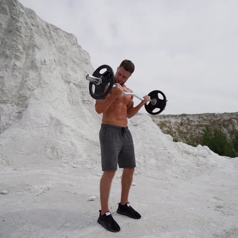 handsome athlete training outdoors. strong shirtless man with muscular body is lifting barbell on the white mountain background in summer. slow motion.