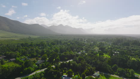 Forwards-fly-above-village-of-suburb-with-houses-surrounded-with-green-vegetation.-Mountain-range-against-sun-in-distance.-South-Africa