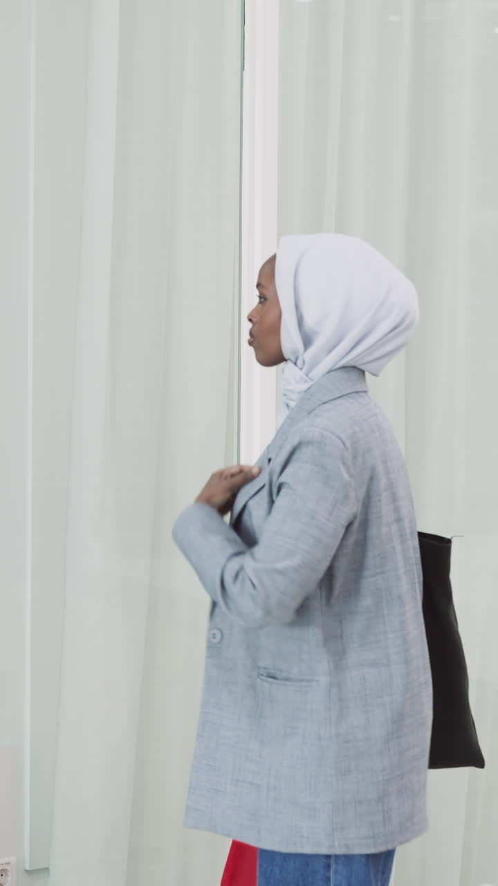 African-American woman adjusts hijab looking at reflection in glass wall slow motion. Muslim lady in jacket visits business center. Arabic tradition